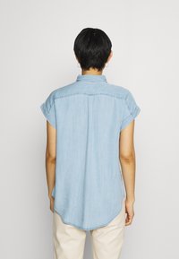 Camicia in denim blu chiaro con maniche corte arrotolate, colletto a punta e orlo curvo. Presenta una texture verticale e una vestibilità casual.