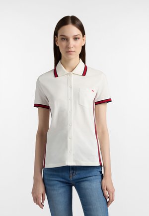 Kvinde med lige brunt hår iført en hvid polo med knapper og røde og marineblå kantbånd, blå jeans, stående foran en ensfarvet baggrund.