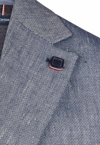 Strukturierter marineblauer Blazer mit einem dezenten diagonalen Muster, versehen mit einer quadratischen Nadel am linken Revers. Futter zeigt das Markenlabel und Streifen.