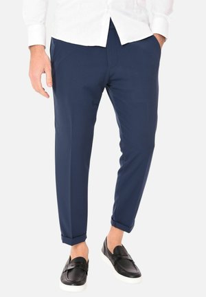 Uomo che indossa pantaloni slim fit blu navy con risvolto, camicia bianca e scarpe slip-on nere con suole bianche, in piedi con le mani parzialmente nelle tasche.