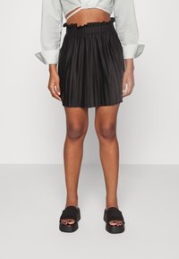 Saia preta plissada com um cós elástico, tecido texturizado e uma borda voluçada na parte de cima. Combinada com sapatos pretos de bico aberto.