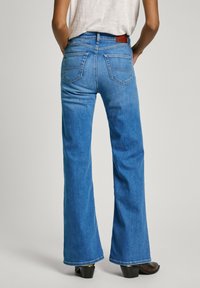 Jean en denim bleu évasé avec une taille mi-haute, deux poches arrière et une patch en cuir à la taille. Le tissu a une texture lisse.