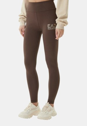 Persona che indossa leggings marroni attillati con il logo EA7 Emporio Armani sulla coscia e sneakers beige chunky, in piedi contro uno sfondo semplice.