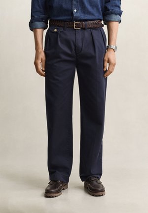 Mann trägt dunkelblaue plissierte Hose, braunen geflochtenen Gürtel, dunkelbraune Lederschuhe und ein Jeanshemd mit hochgekrempelten Ärmeln.