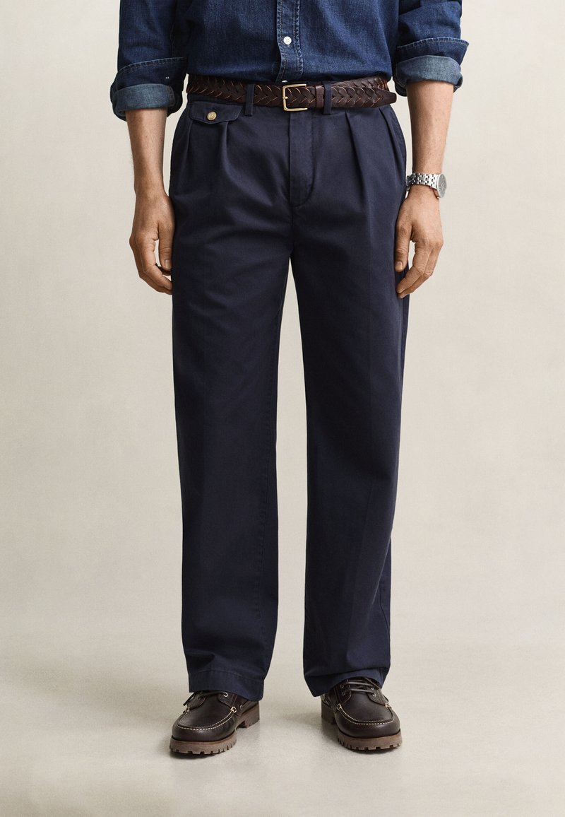 Homme portant un pantalon plissé bleu foncé, une ceinture tressée marron, des chaussures en cuir marron foncé et une chemise en denim avec les manches retroussées.