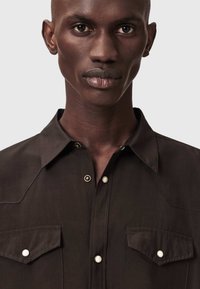Σκούρο καφέ πουκάμισο με κουμπιά, κομψό γιακά, δύο τσέπες στο στήθος και αντίθετα λευκά κουμπιά· λεία υφή υφάσματος και μοντέρνος σχεδιασμός.