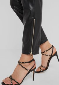 Pantaloni in ecopelle nera con zip laterale, abbinati a sandali neri con tacco alto e fasce con accenti di borchie dorate.