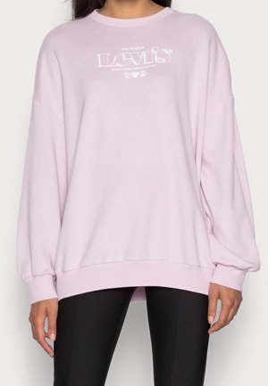 Osoba nosząca jasno różową bluzę Levi's z białym logo, w czarnych dopasowanych spodniach, stojąca z luźno opuszczonymi ramionami.