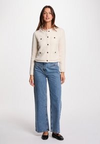 Cardigan texturato color crema con quattro tasche e bottoni marroni, abbinato a jeans a gamba larga blu chiaro e scarpe nere flat.