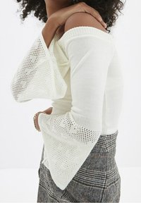 Weißes Oberteil mit schulterfreien Schnitt und strukturierten, ausladenden Ärmeln mit einem Strickmuster. Getragen mit einem gemusterten Karorock.