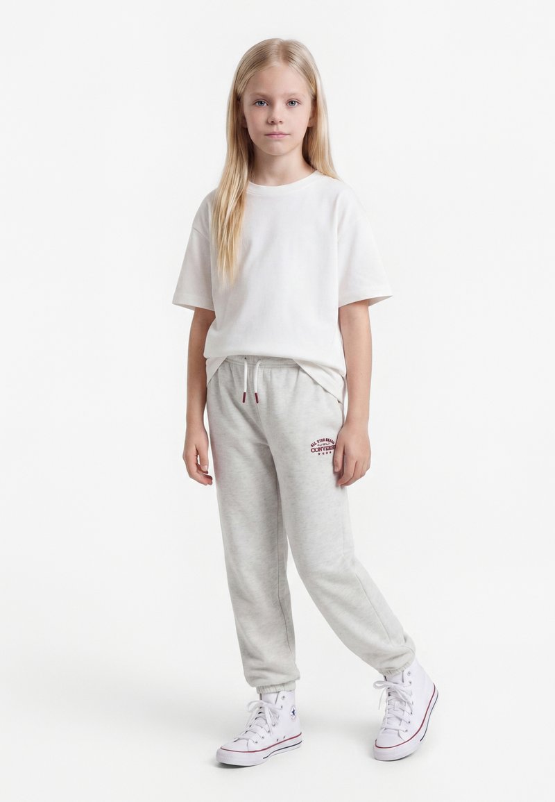 Enfant blond debout, portant un t-shirt blanc, un pantalon de survêtement gris clair avec un logo Converse rouge et des baskets montantes blanches sur fond blanc.