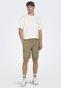 T-shirt blanc à manches courtes avec une poche, associé à un short vert olive avec des ourlets retournés. Le modèle porte des baskets blanches et des chaussettes rayées.