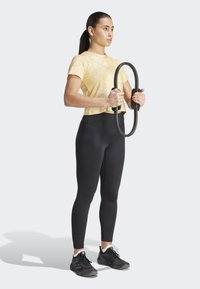 Leggings neri, top corto tie-dye giallo chiaro. Tiene un anello fitness nero. Texture liscia, design atletico, abbigliamento aderente.