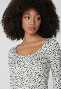 Junge Frau mit lockigem Haar, die ein weißes Oberteil mit kleinen schwarzen Blumenmustern trägt und leicht nach links schaut, vor einem schlichten Hintergrund.