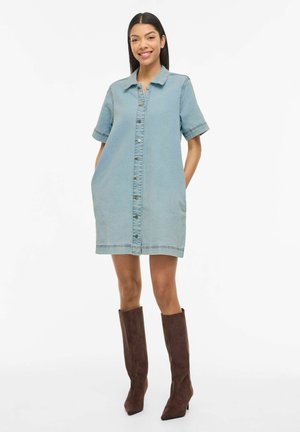 Kvinde iført en lyseblå kortærmet denimkjole med knapper og parret med knæhøje brune ruskindsstøvler, stående og smilende.