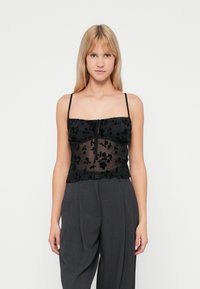 Top in pizzo floreale nero con spalline sottili, centro trasparente e orlo arricciato, abbinato a pantaloni grigi a vita alta.