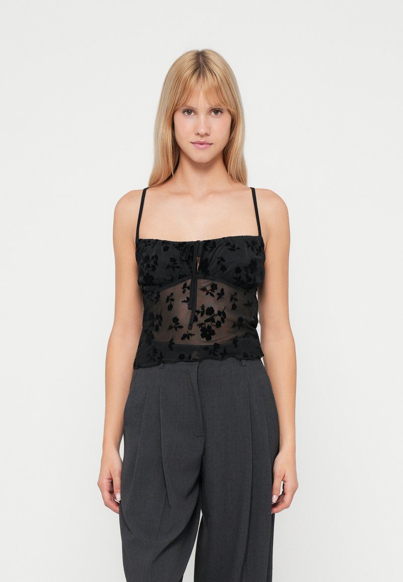 Top in pizzo floreale nero con spalline sottili, centro trasparente e orlo arricciato, abbinato a pantaloni grigi a vita alta.
