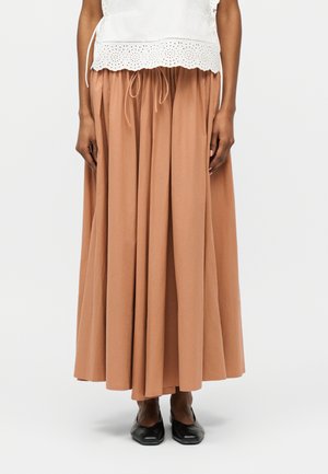 Žena nosi hnedú sukňu širokého strihu s pásom na zaväzovanie, v kombinácii s bielym topom z čipky s očkami a čiernymi špicatými topánkami.