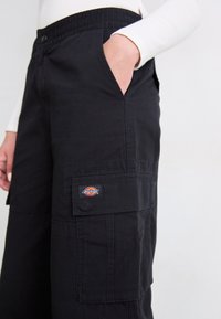 Personne portant un pantalon cargo noir avec une main dans la poche latérale et un logo Dickies visible sur le rabat de la poche cargo.