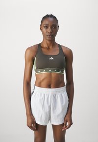 Brun idretts crop top med grønne akenter og logo, kombinert med hvite shorts med svarte sidestriper. Fokuser på stoffets tekstur og passform.