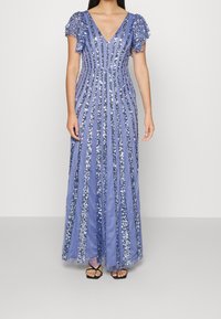 Dlouhé, bezrukávové šaty s výstřihem do V v světle modré barvě s vertikálními flitrovými pruhy. Nabírané krátké rukávy. Rozšířený spodní díl. Nositelný s černými páskovými sandály.