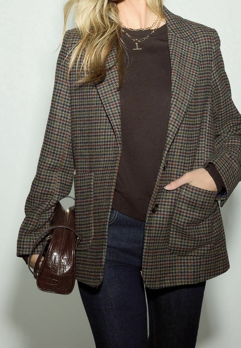 Femme blonde portant un blazer à carreaux sur un top marron et un jean, tenant un sac à main marron texturé, avec une main dans la poche.
