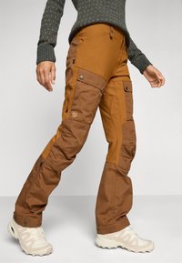 Bruine waterdichte broek met meerdere zakken, een contrasterend paneelontwerp en ritsopeningen bij de boorden, gecombineerd met crèmekleurige schoeisel.