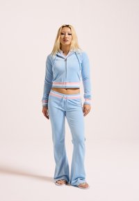 Gaiši zils velūra sporta tērps, kas ietver saīsinātu jaku ar rāvējslēdzēju, rotātu ar rozā un baltām akcentiem, kā arī brīvas formas bikses ar šņorēm vidukļa līmenī.