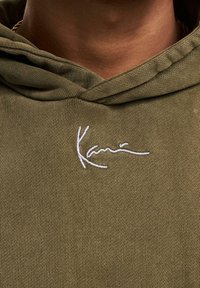 Sweat à capuche vert olive en tissu côtelé, avec un logo signature brodé en blanc sur la poitrine et un cordon de serrage pour la capuche.