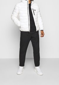 Vit puffjacka med quiltad textur, svart dragkedja och huva, i kombination med svarta byxor och vita sneakers. Minimalistisk design.