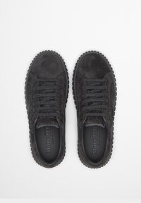 Czarne zamszowe trampki z okrągłymi noskami, płaskimi sznurowadłami i karbowanymi gumowymi podeszwami. Widoczna jest fakturowana powierzchnia i markowe wkładki.