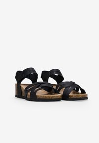 Sandales en cuir noir avec des brides croisées, semelle en liège et talon compensé bas. Semelle texturée pour une meilleure adhérence. Bride de cheville ajustable.
