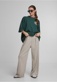 Prekvapujúco veľké zelené tričko s okrúhlym grafickým dizajnom, skombinované s svetlo béžovými širokými nohavicami. Model má na sebe tmavé slnečné okuliare a šľapky.