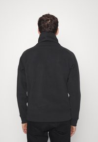Sudadera negra de cuello alto, con puños y dobladillo acanalados. Fabricada en una tela suave con textura lisa. Sin logotipos ni patrones visibles.