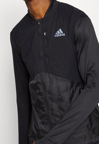 Black athletic jacket with a zip front, textured sleeves, and a smooth body. Features an embroidered blue Adidas logo on the left chest.