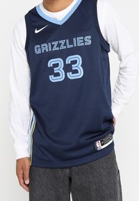 Maillot de basketball bleu marine avec "GRIZZLIES" et le numéro "33" en bleu clair. Présente le logo Nike, des bandes sur les côtés et une étiquette près de l'ourlet. Porté par-dessus une chemise manches longues blanche.