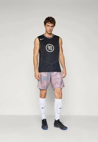 Schwarzes ärmelloses Trikot mit einem kreisförmigen "90"-Logo, kombiniert mit gemusterten grauen Shorts mit roten Wirbeln, weißen Socken und schwarzem Schuhwerk.