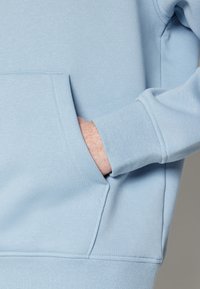 Main dans la poche d'un sweat-shirt bleu clair, montrant le tissu texturé et les détails de couture du poignet.