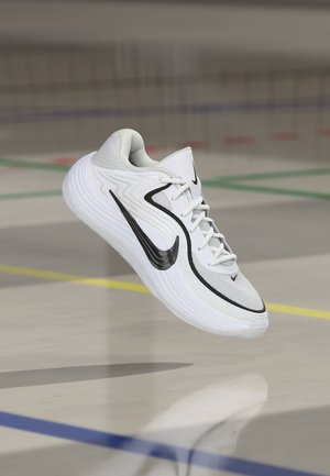 Zapatilla de running blanca de Nike con el logo de la marca en negro, flotando sobre el suelo de un gimnasio marcado con líneas de colores y una red desenfocada al fondo.