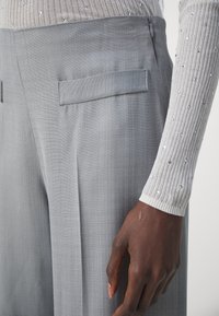 Gros plan sur une main à la peau foncée à côté d'un pantalon sur mesure gris clair avec une poche latérale, assorti d'un haut à manches longues côtelé gris pâle orné de petits strass.