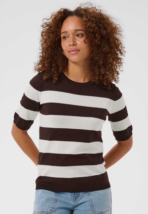 Femme aux cheveux bouclés portant un t-shirt à manches courtes rayé horizontal noir et blanc et un jean bleu clair, debout les mains dans le dos.