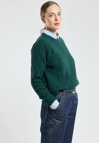Groene gebreide trui met textuurpatronen en kabeldetails, gecombineerd met donkerdenim jeans en een lichtblauw overhemd eronder.