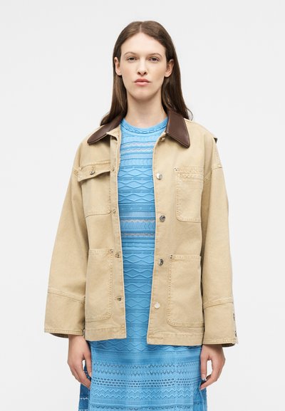 Jeune femme portant une veste beige avec un col marron sur une robe bleu clair texturée, debout devant un fond blanc uni.