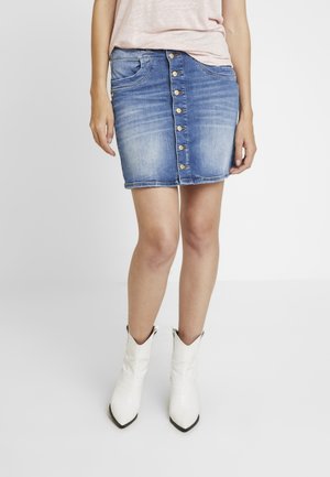 Kvinde iført en blå denim nederdel med knapper foran, lys rosa top og hvide spidse ankelstøvler, stående mod en hvid baggrund.