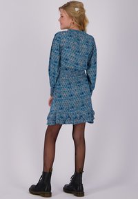 Frau mit blonden Haaren in einem blauen gemusterten langärmeligen Kleid, schwarzen Netzstrumpfhosen und schwarzen Schnürstiefeln, die vor einem schlichten Hintergrund steht.