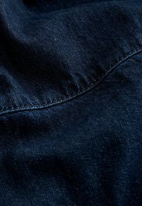 Tissu en denim bleu foncé avec un tissage diagonal visible et une couture horizontale créant de légers plis et une texture.