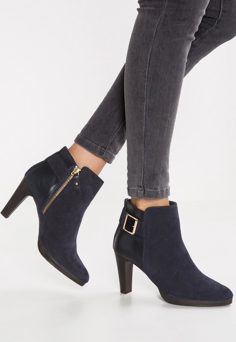 Bottes en daim bleu marine montant jusqu'à la cheville, talons épais, fermeture éclair dorée sur le côté et boucle décorative dorée, portées avec un jean noir retroussé.