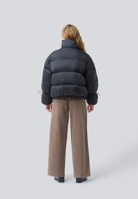 Osoba s dlouhými vlasy, oblečená v černé péřové bundě, širokých taupe kalhotách a černých botách, stojící zády k nám na jednolitém pozadí.