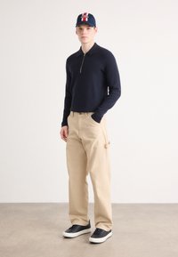 Marinefarbener Zip-Kragen-Pullover, beigefarbene Cargo-Hose mit Seitentasche und schwarze Sneaker mit weißen Sohlen. Männliches Model steht vor einem neutralen Hintergrund.