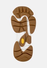 Suola Vibram realizzata in un materiale gommoso marrone con un motivo testurizzato, intersuola bianca e un dettaglio di branding giallo vicino al centro.